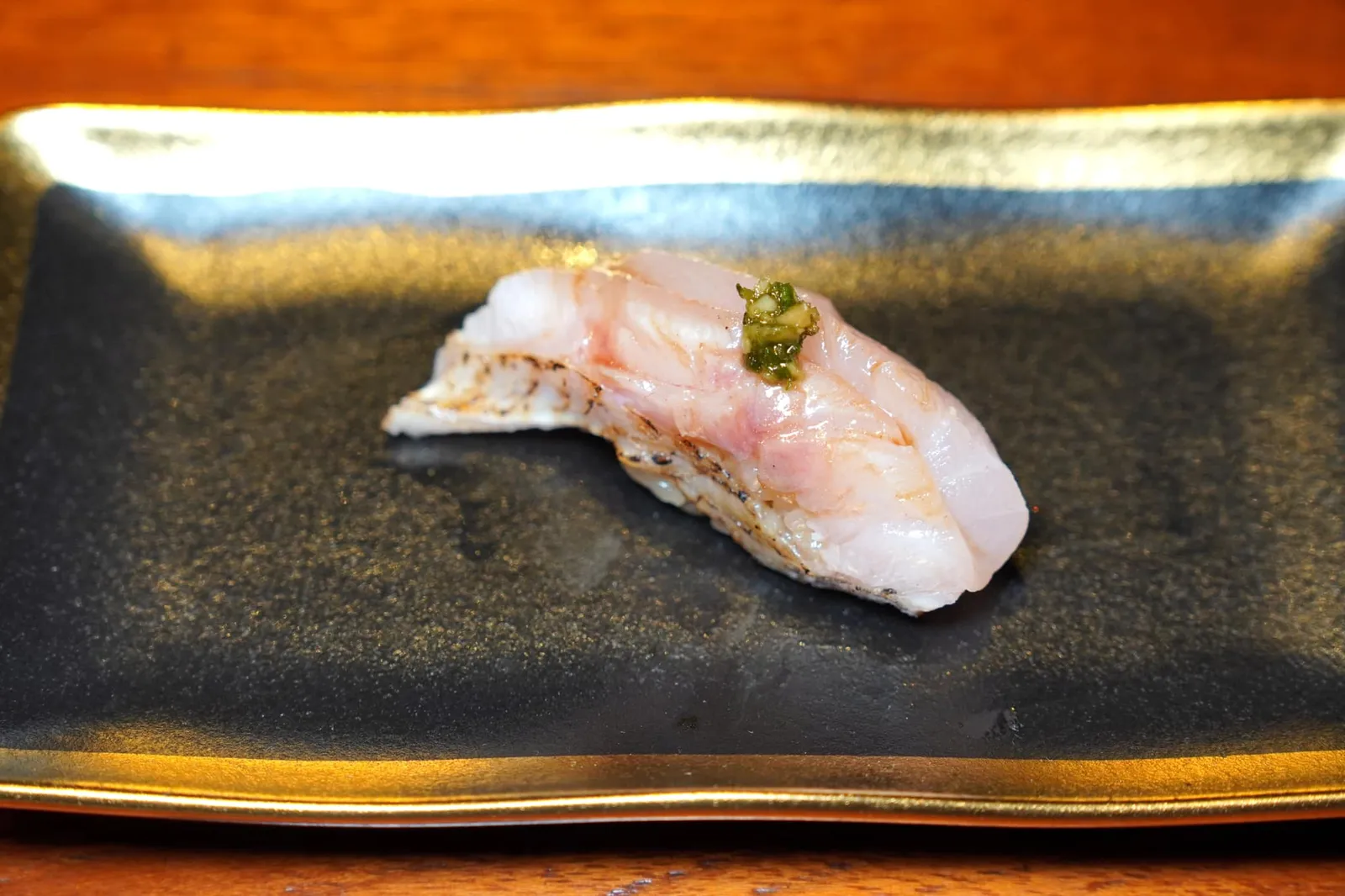 Nodoguro Nirigi Sushi โนโดคุโระหรือปลากะพงคอดำ แม้ตัวปลาจะมีสีชมพูเท่ากันทั้งตัวแต่ชื่อคอดำนั้นมาจากสีในปากของตัวปลานั่นเอง ด้วยความที่เป็นปลาตามฤดูกาลยอดนิยมทำให้ปลาชนิดนี้หารับประทานได้เพียงบางช่วงของปีเท่านั้น ด้านบนทอปด้วยหอมสับช่วยแต่งกลิ่นให้โดดเด่นมากยิ่งขึ้น