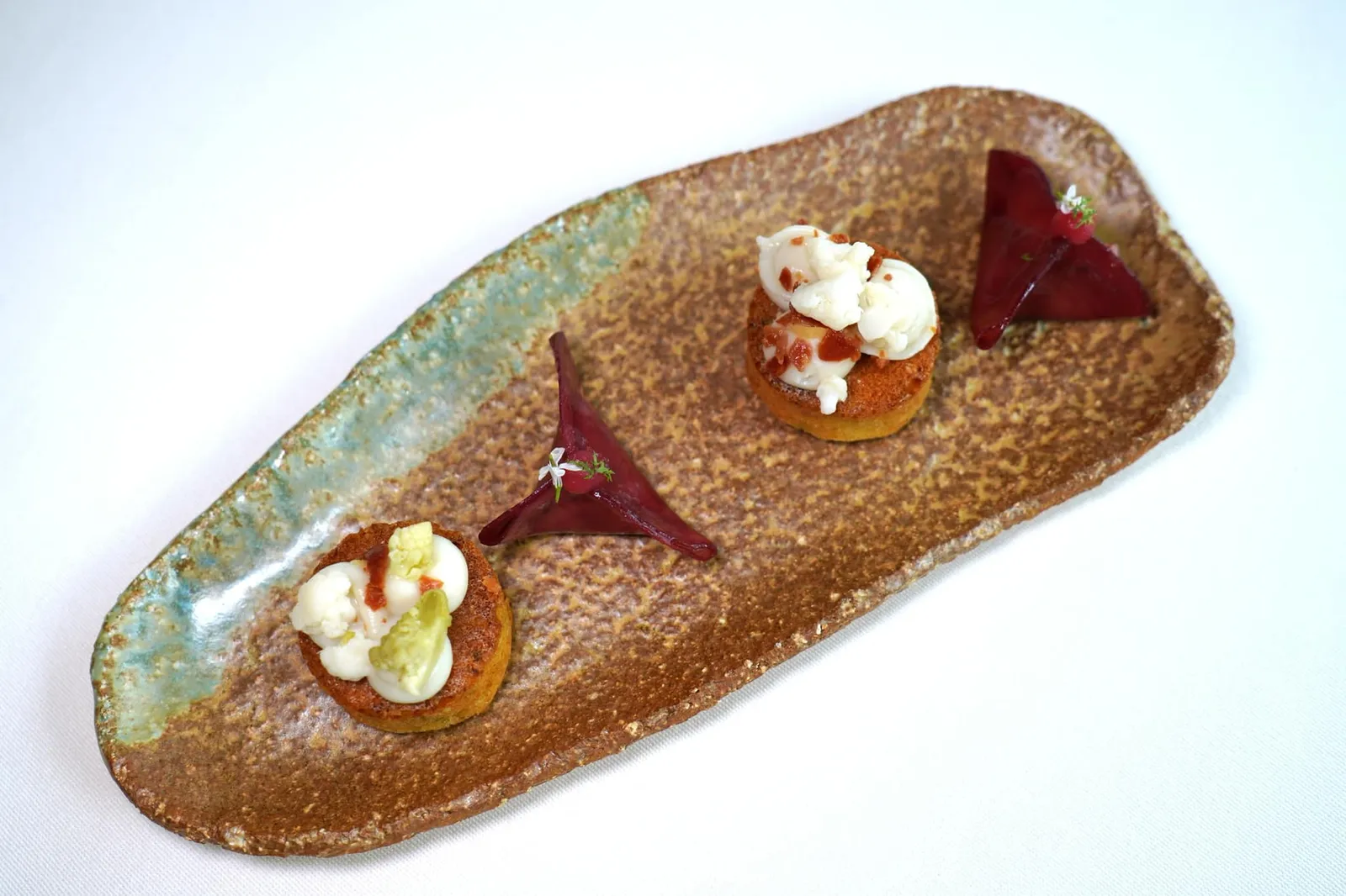 Mises-en-bouche เริ่มต้นด้วยเมนูเรียกน้ำย่อยคือ Beetroot Raviole หรือแผ่นบีทรูทฝานมาเป็นชิ้นบางกรอบแล้วห่อเป็นรูปเกี๊ยว ด้านในสอดไส้ด้วยเนื้อปลาเทราต์ออร์แกนิคจาก Pisciculture De Crisenon ฟาร์มปลาชื่อดังแห่งแคว้น Burgundy คำนี้ให้รสชาติโดยรวมออกไปทางเปรี้ยวแต่กลับไม่มีกลิ่นดินของบีทรูทติดมามากจนเกินไปช่วยกระตุ้นความอยากอาหารได้ดี (17/20) ข้างกันคือ Cauliflower Cake นุ่ม ๆ หอม ๆ ยังมีกะหล่ำดอกที่ถูกหั่นเป็นชิ้นจิ๋ว ๆ ช่วยบาลานซ์ความเปรี้ยวจากบีทรูทคำก่อนหน้า (17/20)