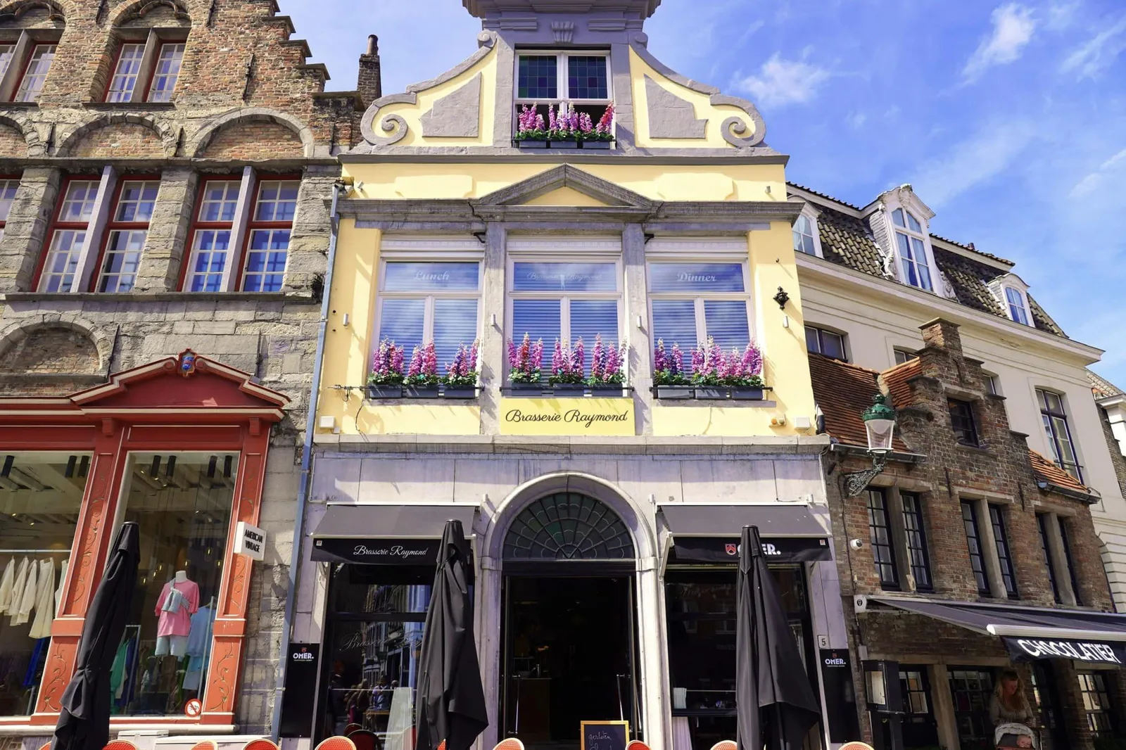 ด้านหน้ามองเห็นเป็นอาคารสีเหลืองอ่อนที่มีลักษณะสถาปัตยกรรมแบบบ้านเก่าของนคร Brugges