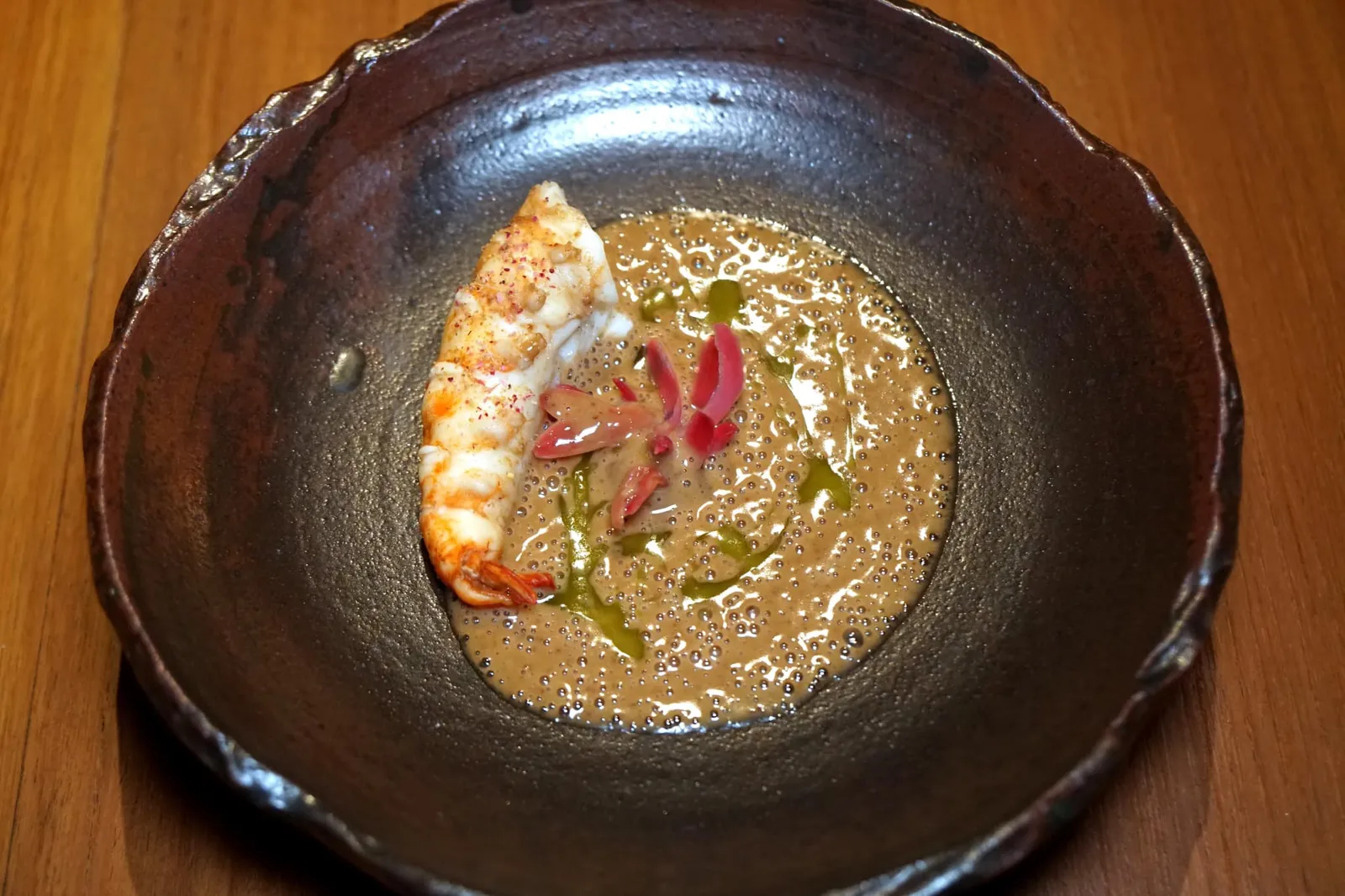 Chef's first trip to Surat Thani River prawn and fermented soy bean คอร์สนี้เป็นจานที่เชฟได้รับแรงบันดาลใจมาจากตอนที่ไปจังหวัดสุราษฎร์ธานีเป็นครั้งเเรก จึงได้นำ “กุ้งเเม่น้ำธรรมชาติ” จับจากแม่น้ำตาปีมาเป็นวัตถุดิบหลัก เริ่มจากการนำไปแช่ในสาโทไทยเป็นระยะเวลา 1 ชั่วโมงเพื่อให้ความหวานของข้าวหมักเเทรกซึมเข้าไปในเนื้อกุ้ง จากนั้นนำไป Poached ในซอสสีน้ำตาลทำมาจากถั่วเน่าเดือด เนื้อกุ้งนุ่ม เด้ง สุกกำลังดี โรยด้วยผงสีเเดงด้านบนทำมาจากดอกดาหลา เชฟนำซอสถั่วเน่าซึ่งมีกลิ่นเเละรสชาติเฉพาะตัวเสิร์ฟมาข้างกัน สีเขียวด้านบนคือ Vegetable Oil เเหวกดูในตัวซอสจะพบกับดอกดาหลาที่ผ่านการดองจนมีรสชาติเฉพาะตัว ก่อนเสิร์ฟเชฟนำกุ้งเเม่น้ำไปรมควันกับไม้ลำไยอีกทีเพื่อเพิ่มความหอมให้เด่นชัดมากยิ่งขึ้น (16/20)