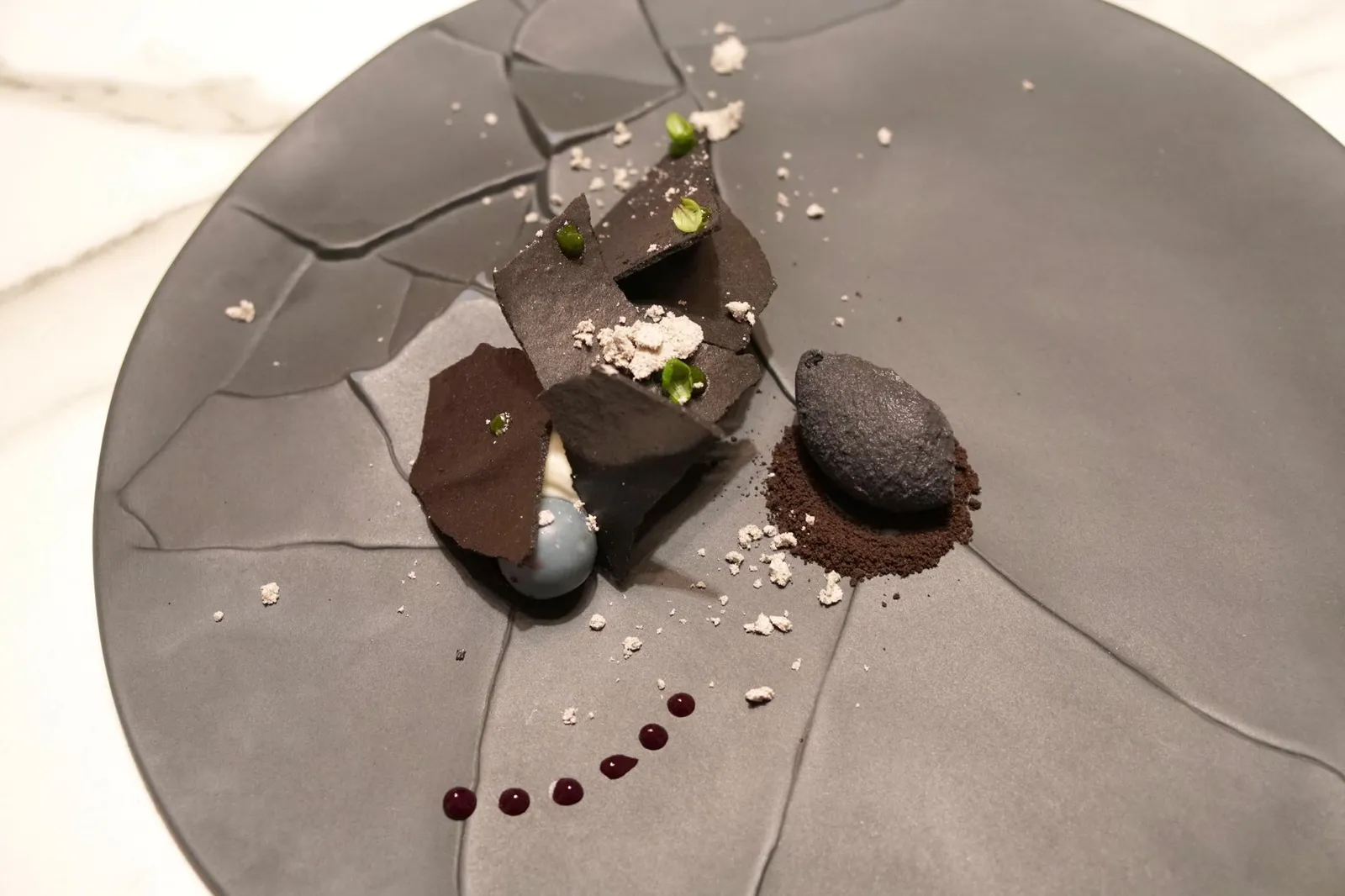 COCONUT GANACHE, BLACK GARLIC, BLACKCURRANT BUBBLEGUM, BASIL& LIQOURICE... COCONUT-YUZU ASHES ICE CREAM. ขนมหวานจานเเรกเสิร์ฟมาในจานที่ดูคล้ายจานแตก เป็นกานาชสีขาวทำจากมะพร้าวเเอบซ่อนอยู่ด้านล่างของจาน ทานกับขนมหมากฝรั่งบลูเบอร์รี่ 2-3 ลูก แผ่นกรอบทำมาจากกระเทียมดำและ liqourice rice ฝั่งขวาคือไอศกรีมสีดำสนิททำมาจากเถ้าของมะพร้าวเเละส้มยูซู เถ้ากรอบด้านล่างรสชาติคล้ายบิสกิตโอรีโอ้ พนักงานเเนะนำให้เริ่มทานตัวหมากฝรั่งก่อน มีกลิ่นหอม เเละจะเเตกระเบิดออกในปากเวลาทาน ไอศกรีมมีรสและกลิ่นของส้มยูซูชัดเจนผสมกับกลิ่นมะพร้าวเบาๆ เข้ากันอย่างลงตัว ถือเป็นขนมที่มีองค์ประกอบซับซ้อน มีลูกเล่นในตัวเอง และมีเอกลักษณ์เฉพาะตัวสูงมากจริงๆ ... ความแปลกอย่างเดียวของจานนี้คือพนักงานให้ช้อนมาอย่างเดียวทำให้ตักทานยากไปหน่อย (20+/20)