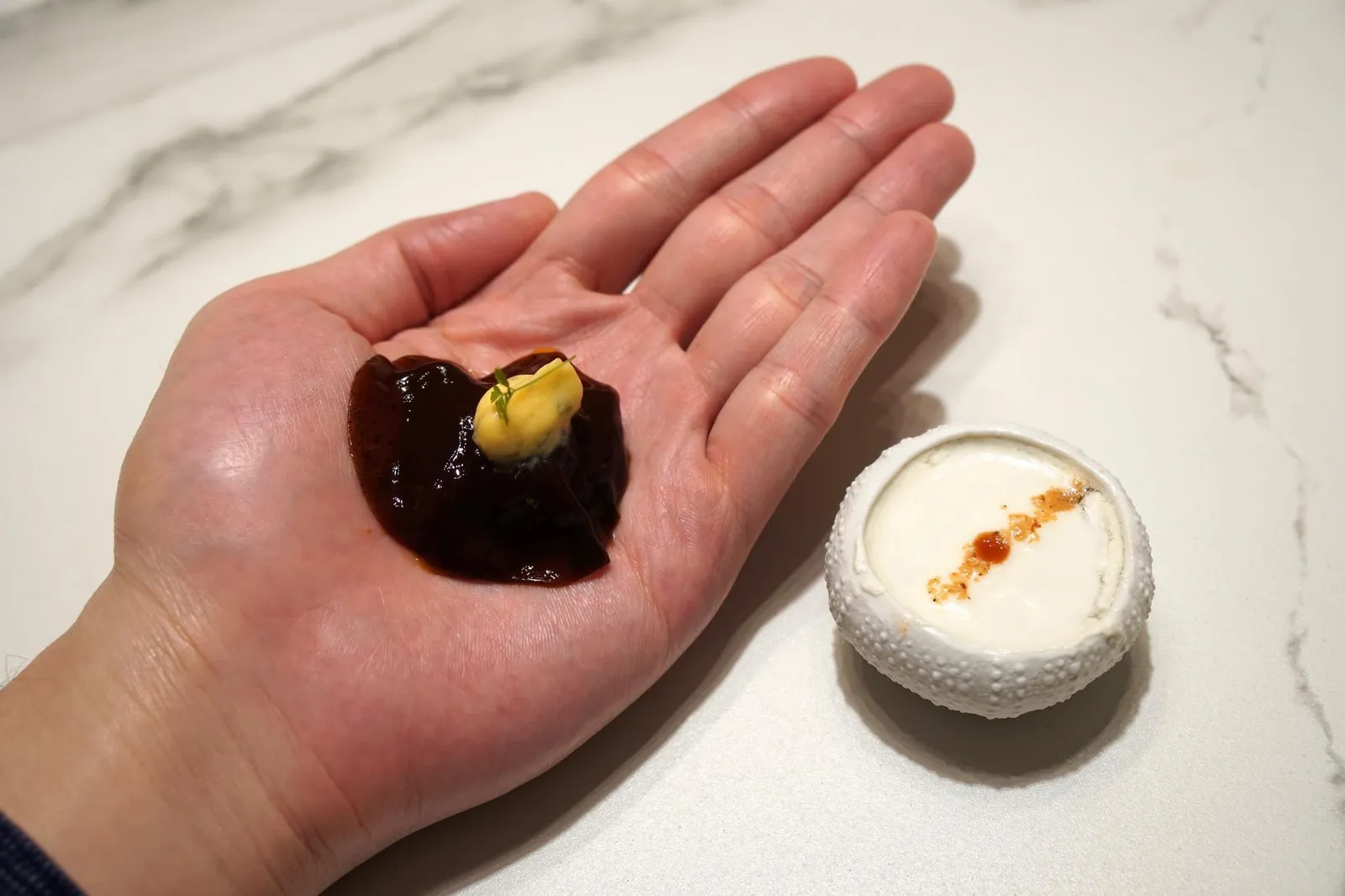 TRIP TO TSUKIJI MARKET... "SEA URCHIN ON THE HAND" WITH FINGER LIME. A VEIL OF "LIQOURICE-BLACK GARLIC" JAPANESE BEARNAISE SAUCE AND BERGAMOT SCENT. สีส้มข้างบนคือ Bearnaise sauce สไตล์ญี่ปุ่นทำจากเนย วางใบผักชีไว้ข้างบน ปิดท้ายด้วยการฉีดน้ำหอมกลิ่น Bergamot ตกแต่งเล็กน้อยโดยการทอปใบผักชีลงไปข้างบน ตอนทานเข้าไปรสชาติแรกที่สัมผัสได้คือความสดของไข่หอยเม่นเย็นๆ เนื้อสัมผัสเนียน ละมุน ครีมมี่สุดๆ ตัดรสเปรี้ยวด้วยมะนาวคาเวียร์ที่แตกออกในปาก วัตถุดิบทั้งสองอย่างดูเหมือนจะมีรสชาติที่เเตกต่างกันเเต่กลับถูกเชื่อมด้วยซอส Bearnaise ที่ทำจากเนยได้อย่างลงตัว กลิ่นของเจลพริกไทยกับกลิ่นมะกรูดเสริมกันได้พอเหมาะพอดี (20/20)