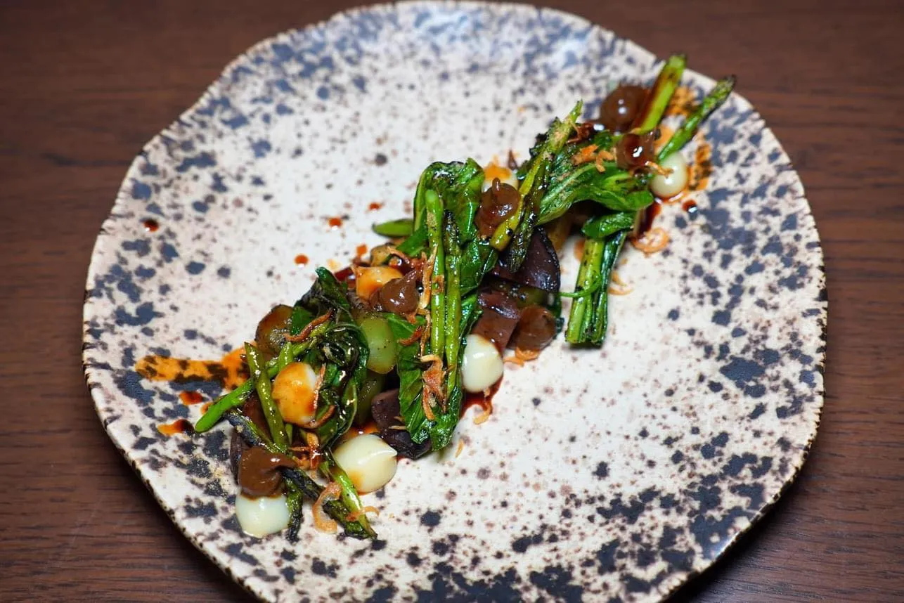 “Vegetable of the Day, ผักตามฤดูกาล” ผัดผักตามฤดูกาลประกอบไปด้วยคะน้า หน่อไม้ฝรั่งที่ปลูกในประเทศไทย เห็ดตับเต่า และผักวอสุ่นจากดอยแม่สะลองจับคู่กับพูเรกระเทียมสีขาวที่ทางร้านทำเอง ยังมีซอสคาราเมลสีน้ำตาลที่เตรียมขึ้นมาจากส่วนที่เหลือจากหอยนางรม จากนั้นจบด้วยการเทราดน้ำมันหอยสูตรพิเศษของทางร้ายลงไปด้านบน (14/20)