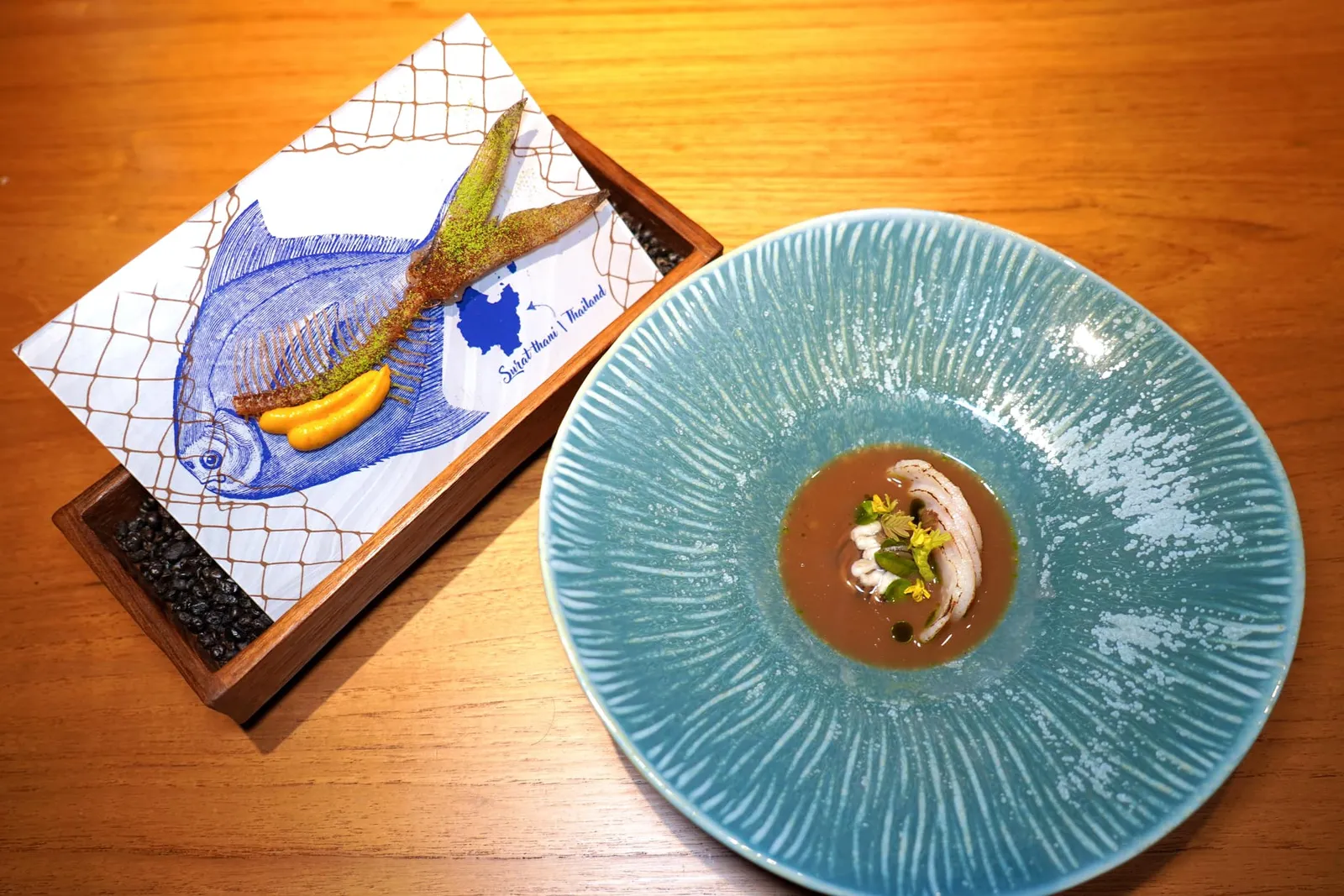 LOCAL WISDOMSNAKEHEAD FISH | ANT EGGS | GRILLED AND GRILLED SHEATFISHES | FISH ROE | YANANG LEAVES | BABY BAMBOO SHOOTS | LOCAL THAI FERMENTED FISHเชฟนำเสนอต้มโคล้งรูปแบบใหม่โดยใช้ปลาจาระเม็ดขาวที่ผ่านการ Poached ในน้ำตาลสมุนไพรที่มีความหอมของปลาแห้ง ด้านล่างรองด้วยน้ำมันใบกะเพรา ข้าง ๆ กันคือหน่อไม้คลุกเคล้ากับเจลใบย่านาง จากนั้นท็อปด้วยเห็ดฝานเป็นชิ้นบาง ๆ ย่างจนสุกรวมไปถึงเจลปลาร้าและไข่มดแดง จากนั้นที่โต๊ะเชฟจะเสิร์ฟน้ำซุปที่เตรียมมาจากปลาเนื้ออ่อนที่ผ่านการรมควันกับไม้ลำไยปรุงรสชาติด้วยใบมะขามอ่อน อีกฝั่งหนึ่งคือก้างปลาจาระเม็ดแห้งทอดกรอบเสิร์ฟมาคู่กันกับผมใบมะกรูดและผงพริกคั่ว นอกจากนี้ยังมีซอสพริกเหลืองย่างผสมไข่ปลาใส่มาอีกด้วย (13/20)