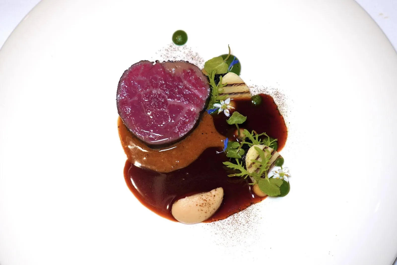 NEW ZEALAND WAGYU BEEFBeef Fat, Root Vegetables, Cumin Leavesเนื้อผัดใบยี่หร่าจานนนี้ได้จากการนำเนื้อวัววากิวจากประเทศนิวซีแลนด์ไปย่างบนเตาถ่าน ระหว่างนั้นค่อย ๆ ทาด้วยไขมันวัวเรื่อย ๆ ตลอดช่วงเวลาที่ย่าง ข้าง ๆ คือเพียวเรสีเหลือง่อนของหัวหอมกงฟี เพียวเรสีเขียวของใบยี่หร่าและสมุนไพร ชิ้นยอดมะพร้าวที่ Poached กับกะทิต่อด้วยการย่างและรมควัน ชิ้นสีดำคือมันขี้หนูที่ผ่านการกงฟีและย่างย่าง ก่อนจะเกลซด้วยซอสที่ผสมผสานหัวหอมไหม้และกระเทียมต้นไหม้เข้าด้วยกัน ซอส 2 ชนิดก็มีความน่าสนใจเช่นกันโดยซอสสีอ่อนกว่าเชฟใช้ชื่อเรียกว่า Beef Fat Sabayon เนื่องจากใช้ไขมันวัวแทนที่เนยปกติ ส่วนซอสสีเข้มคือซอสไวน์แดงที่ Slow-cooked กับสมุนไพรและเครื่องเทศ จากนั้น Infused ด้วยใบยี่หร่าสดเพื่อเพิ่มความร้อนเล็กน้อย (15/20)
