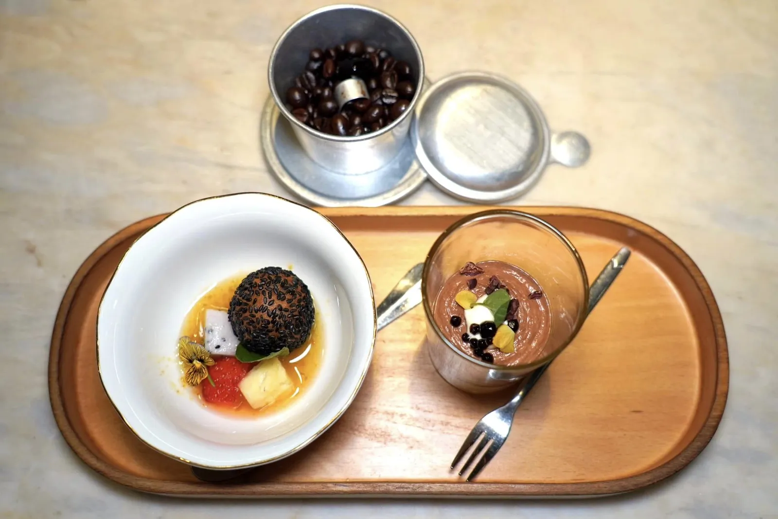 BÁNH CAMRice mochi ball, Marou 70% chocolate, ginger calamansi sauceBáhn Cam เป็นขนมที่ขายกันตามร้านอาหารริมทางโดยมีต้นกำเนิดมาจากประเทศจีน แป้งข้าวเหนียวกรอบนอก นุ่มใน ด้านนอกเชฟคลุกมากับงาดำ เชฟแทนที่ถั่วเหลืองรสหวานด้วยกานาซดาร์คช็อกโกแลต Marou จากจังหวัด Tien Giang ในแถบ Mekong River Delta (13/20)CÀ PHÊ SỮADark chocolate mousse, Robusta coffee, Valrhona milk chocolateCà Phê Sữa เป็นขนมท้องถิ่นที่ผสมผสานกาแฟเวียดนามเข้ากันกับนมข้น เชฟเตรียมมาในลักษณะของมูสกาแฟที่มีรสชาติเข้มข้นจากเมล็ดกาแฟ Robusta ท้องถิ่นคั่วผสมผสานกันกับดาร์คช็อกโกแลต Marou นอกจากนี้เชฟยังแทนที่นมข้นหวานด้วยองค์ประกอบของไวท์ช็อกโกแลตจาก Valrhona เช่นกัน ส่วนในถ้วยข้าง ๆ กันคือเจลลี่กาแฟนุ่มหนึบ (13/20)