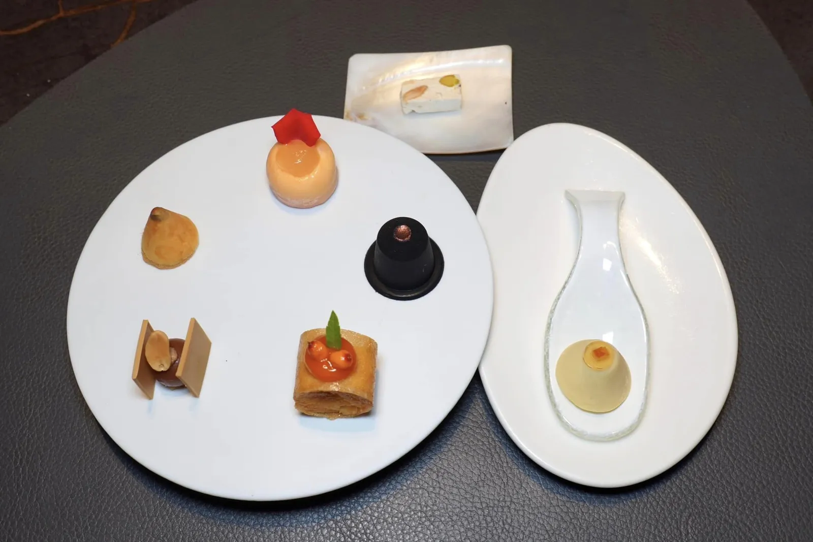 Petits foursขนมชิ้นจิ๋วเริ่มต้นจาก 12 นาฬิกาแล้ววนไปตามเข็มคือ Apricot Parfait with Pineapple Gel, Pressed Macchiato Cup Praline, Rum Cheesecake, Peanut-Caramel Fudge และ Cornetto with Eggnog Cream and Chocolate ด้านบนคือ Montélimar Nougat ส่วนในช้อนคือ Chai Latte with Grand Marnier Cream; Orange and White Chocolate