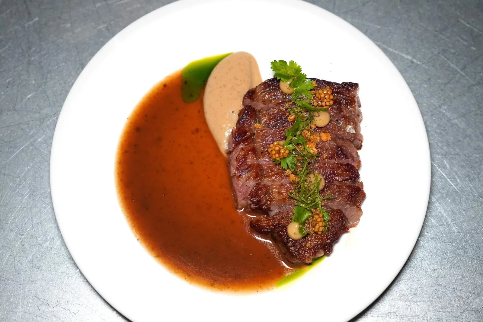 WAGYU STRIPLOIN (690++)เนื้อวัววากิวส่วนสันนอกเสิร์ฟมาที่ความสุกมีเดียมพอเหมาะ จากนั้นราดด้วยซอสที่ได้รับแรงบันดาลใจมาจากเกาเหลาเนื้อแบบไทย ด้านบนคือเพียวเรกระเทียมและสมุนไพรต่าง ๆ