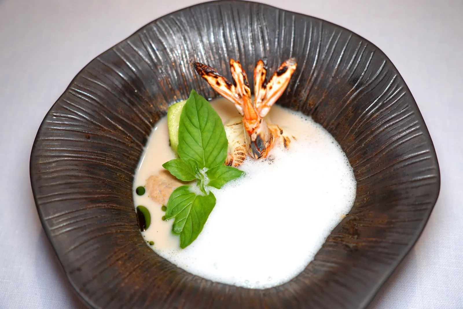 Authentic Kang Liang Soup with River PrawnBanana Prawn | White Peppercorn | Shrimp Paste | Lemon Basil Foam“แกงเลียง” สูตรพื้นบ้านที่มีความหอม มัน ครีมมี่ และอุมามิจากกะปิเคยชั้นดีผสมผสานกับโฟมเลมอนเบซิลและน้ำมันผักชี ในจานยังมีซูคินี่ เห็ดฟาง และฟักทองน้ำเต้า ส่วนโปรตีนหลักคือกุ้งย่างถ่านที่จะช่วยเพิ่มเติมกลิ่นและความหอมลงไปในน้ำแกงนั่นเอง (14/20)